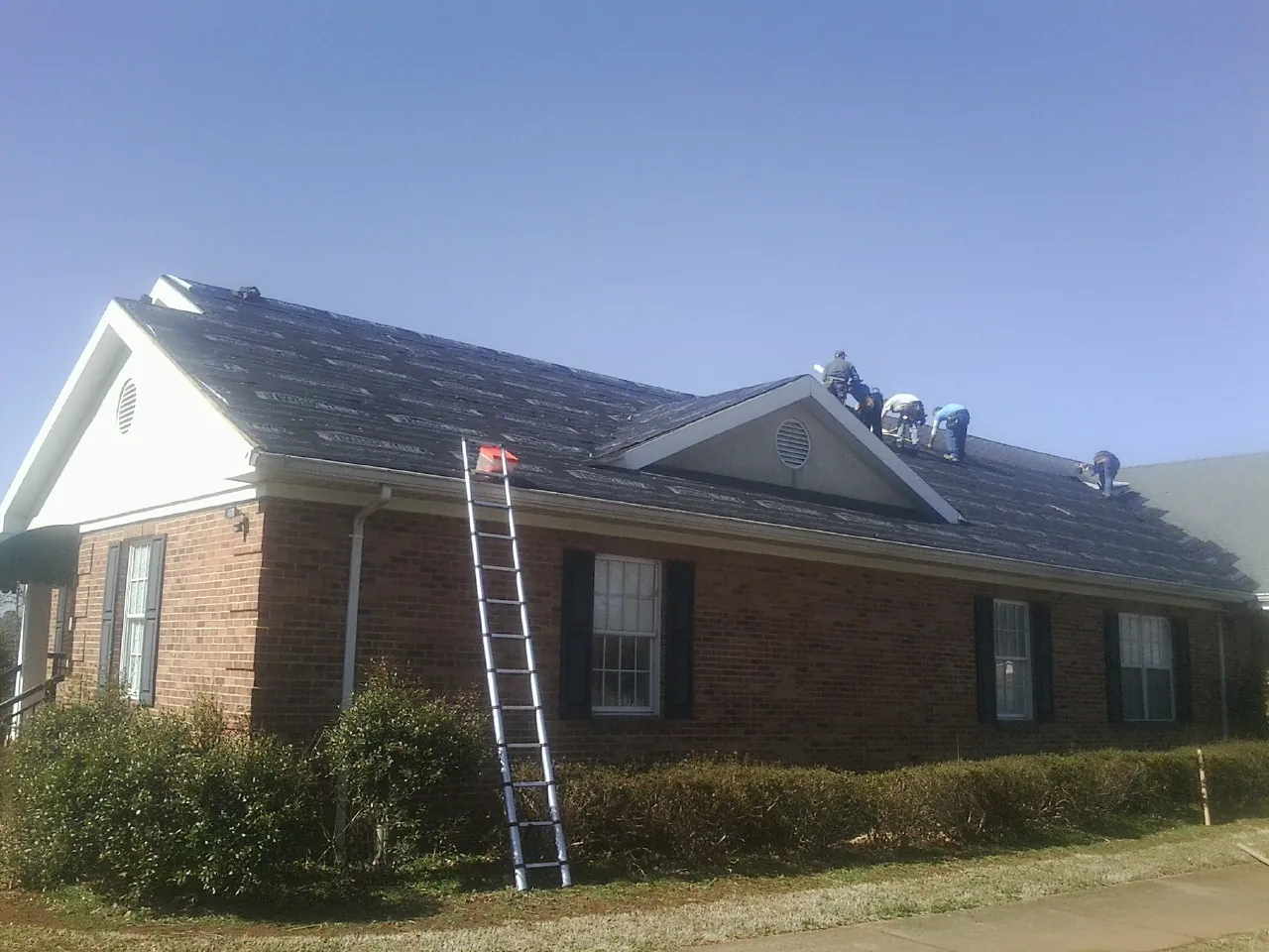 Crew tearing shingles off a brick ranch roof with a ladder set — Metal Roof Repair work by our crew in Lamar